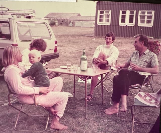 A family sitting around a camping table in front of one of the old wooden army huts