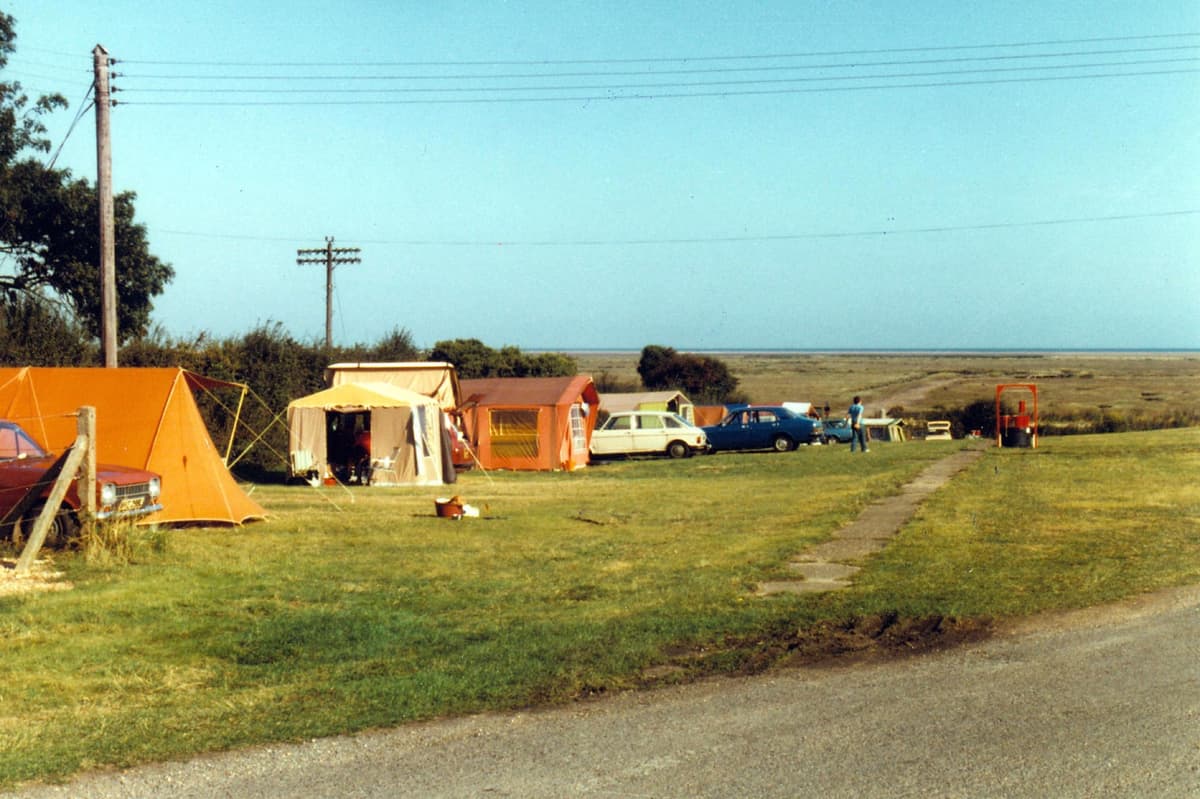 A wide view of the site in 1979, tents and cars pitched on the grass beside the marsh
